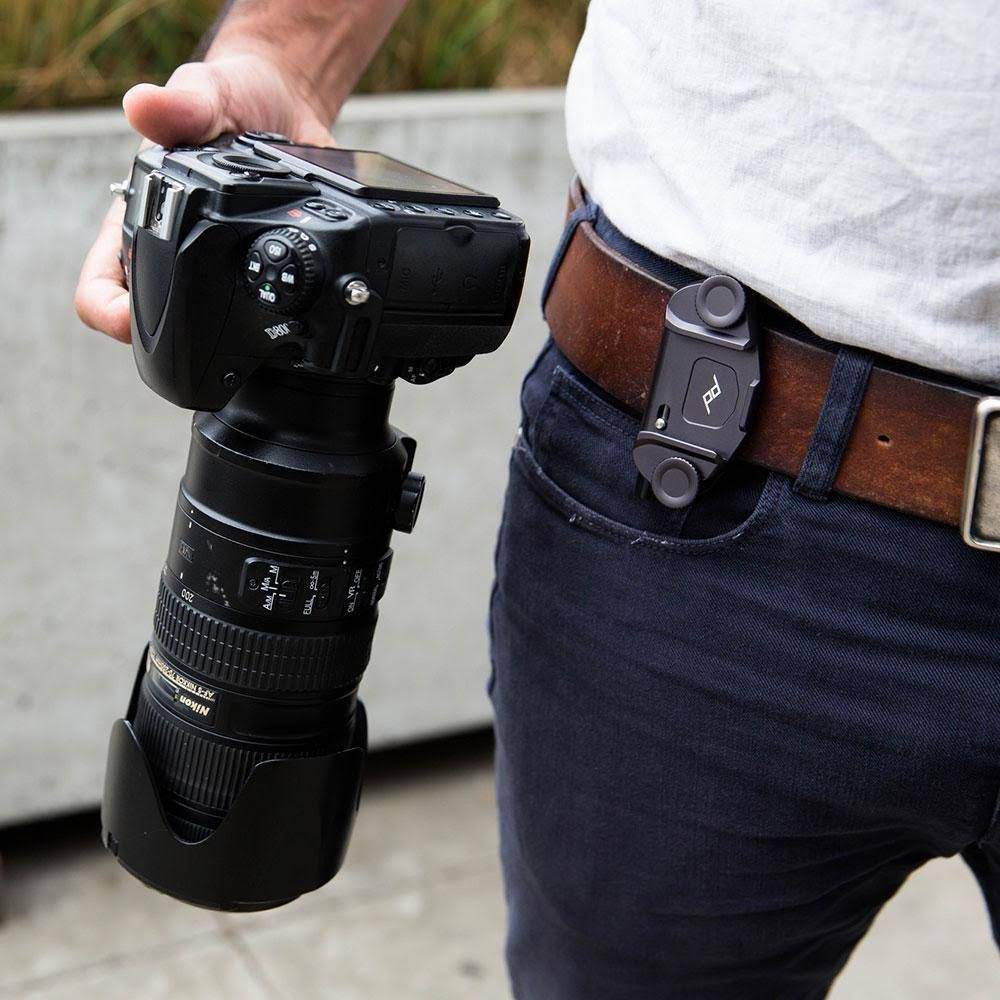 Osoba trzyma czarny aparat fotograficzny z dużym obiektywem, przypięty do paska.
