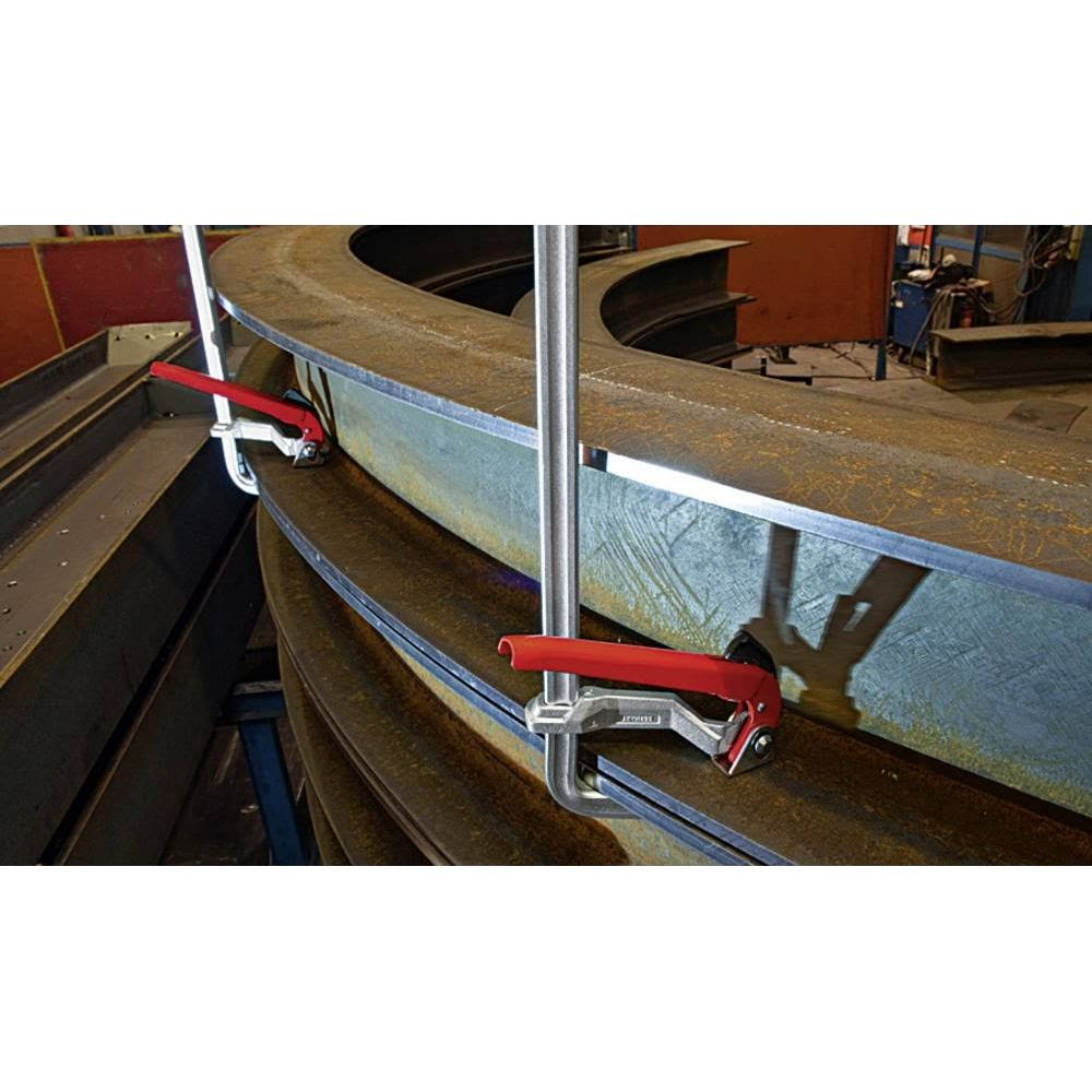 Zwei rote Klemmen halten gebogene Stahlträger im Werkstattbereich.