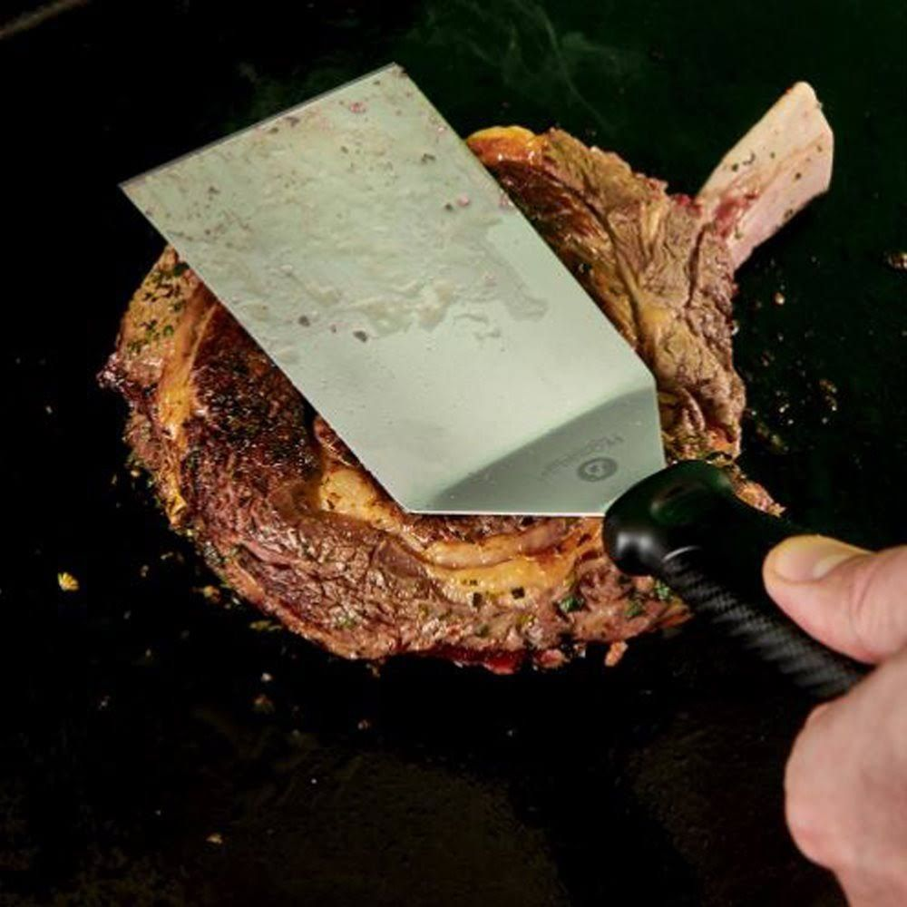 Ein Steak wird mit einem Metallspatel auf einer schwarzen Oberfläche gewendet.