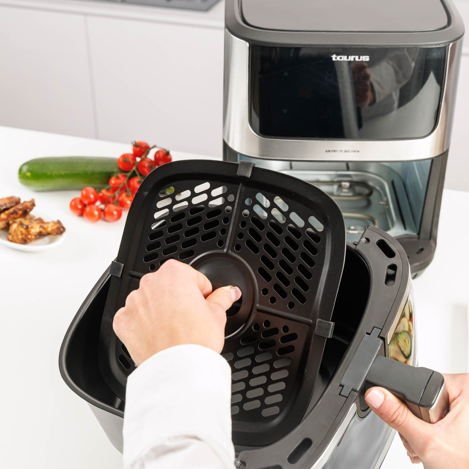 Una mano retira la cesta de una freidora de aire, con ingredientes cerca.