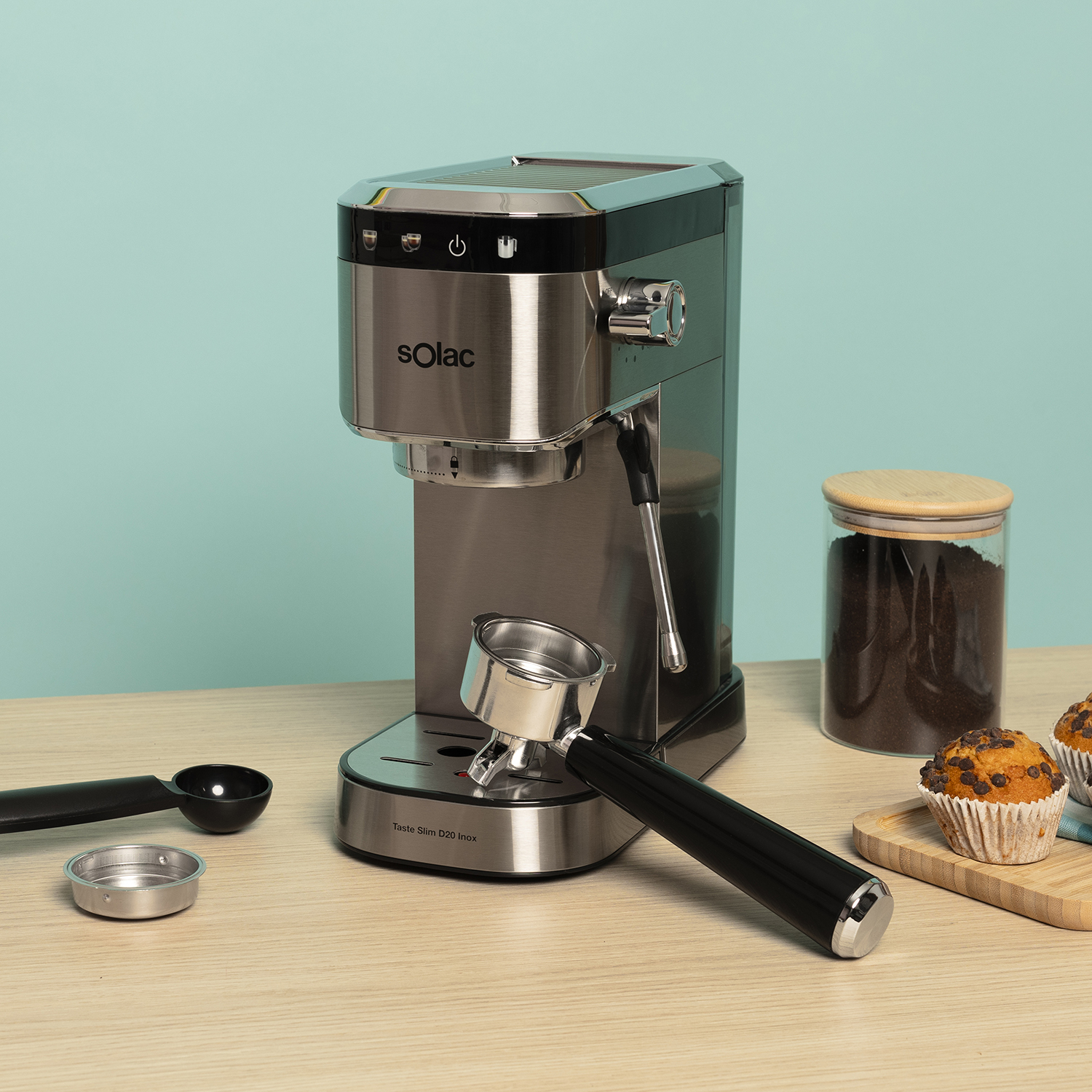 Una cafetera espresso plateada con mango negro sobre mesa de madera. Al lado, cuchara, filtro y muffins.