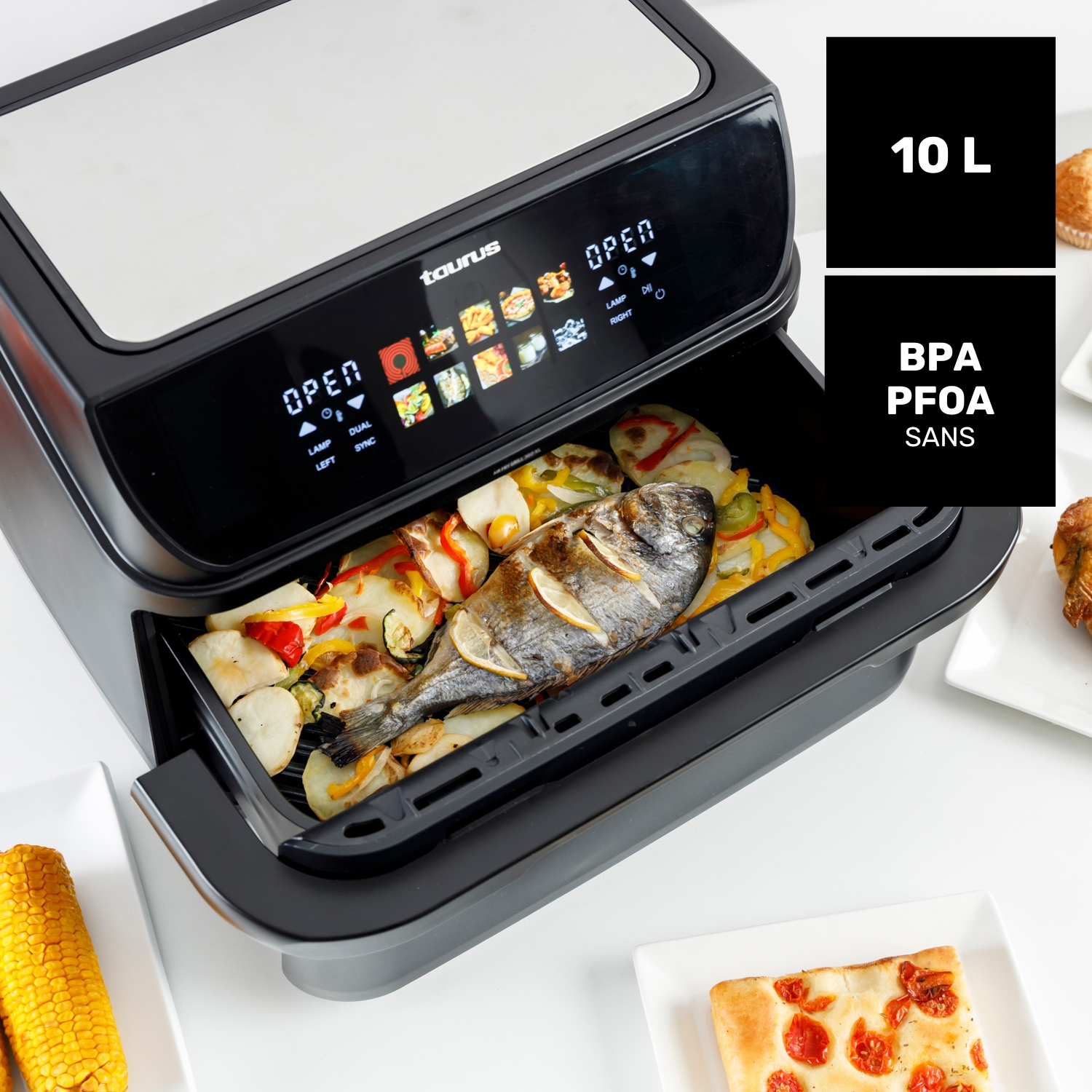 Una freidora de aire-grill negra con pescado y verduras asados, mazorca de maíz y focaccia.