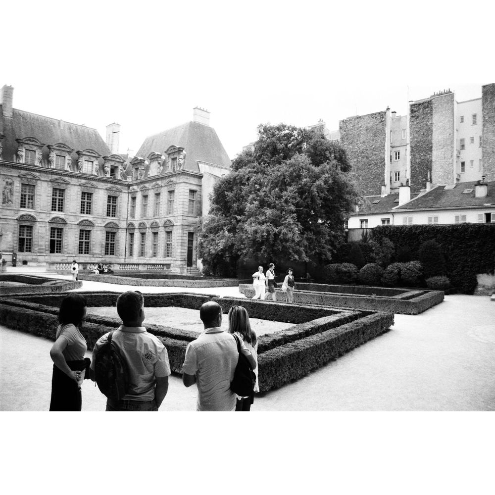 Ludzie w ogrodzie przed zabytkowym budynkiem z drzewami.