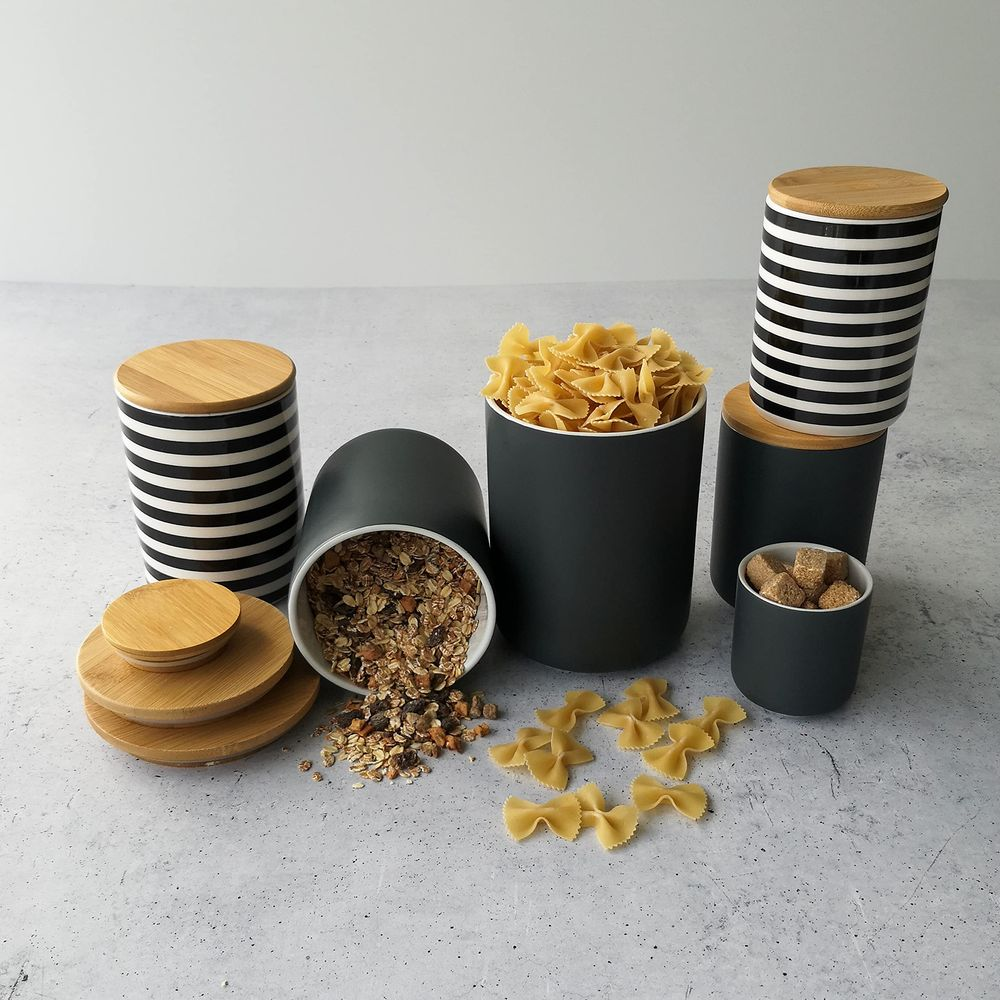 Dunkelgraue Vorratsgläser mit Holzdeckeln, gefüllt mit Müsli, Nudeln und Zuckerwürfeln.