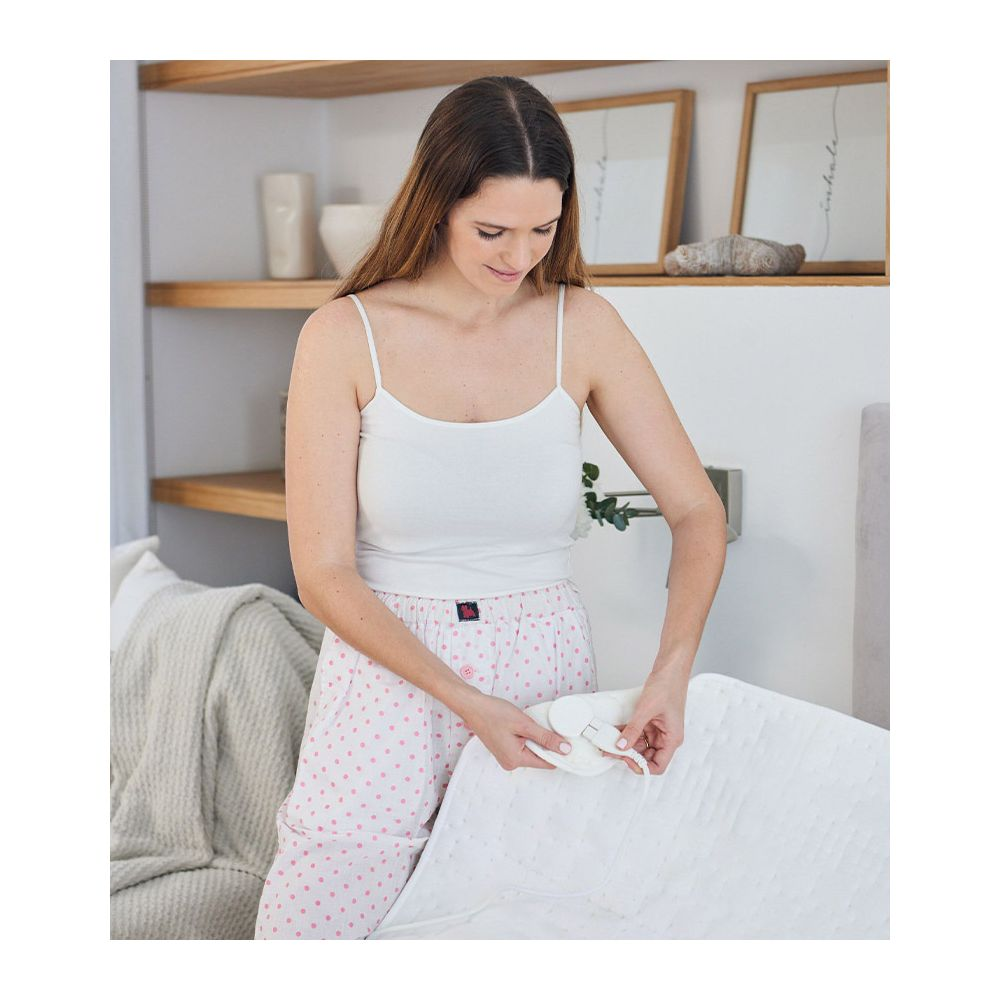 Eine Frau in weißem Top und rosa gepunkteter Hose hält ein weißes Heizkissen.