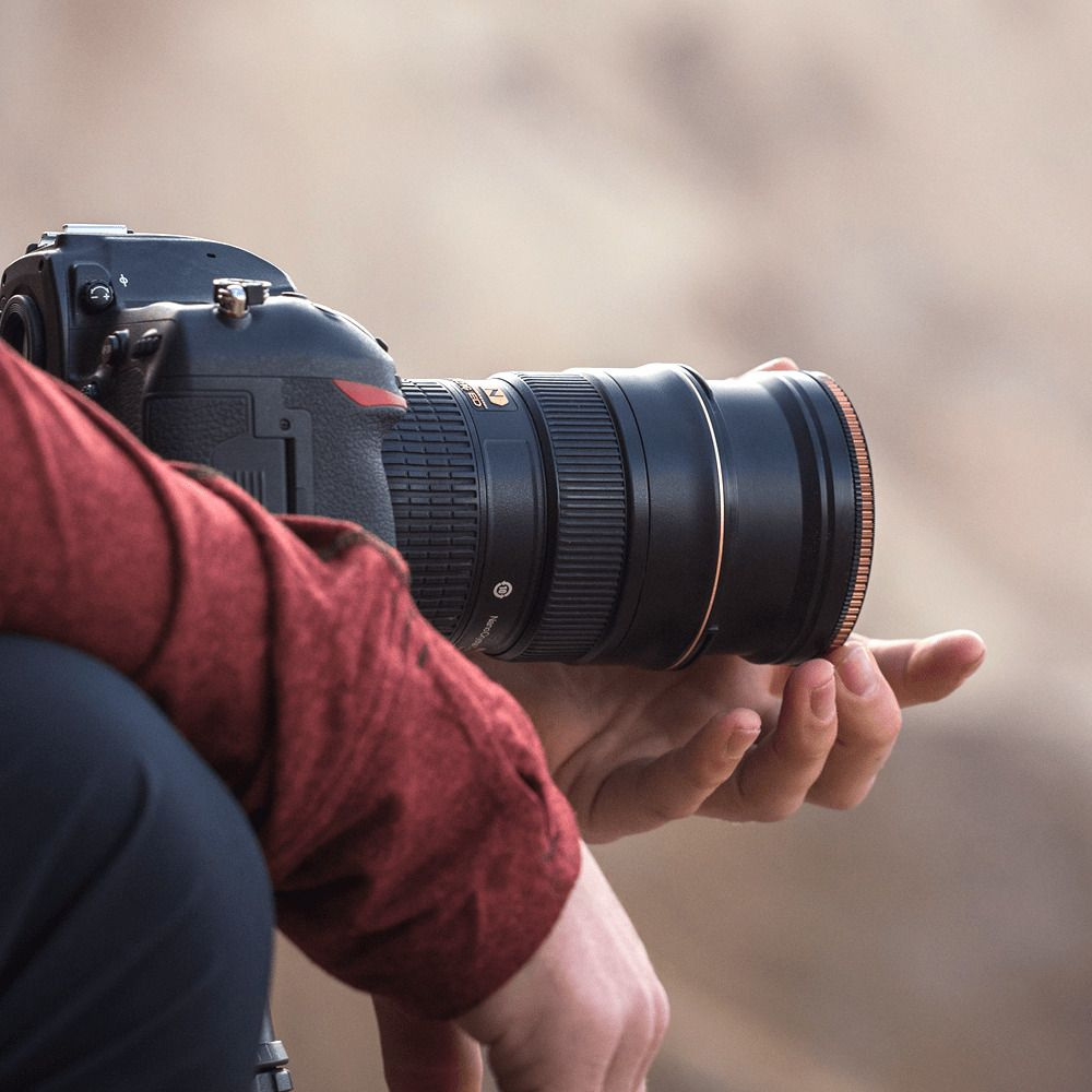 Een persoon in een rood shirt houdt een zwarte DSLR-camera met grote lens vast.