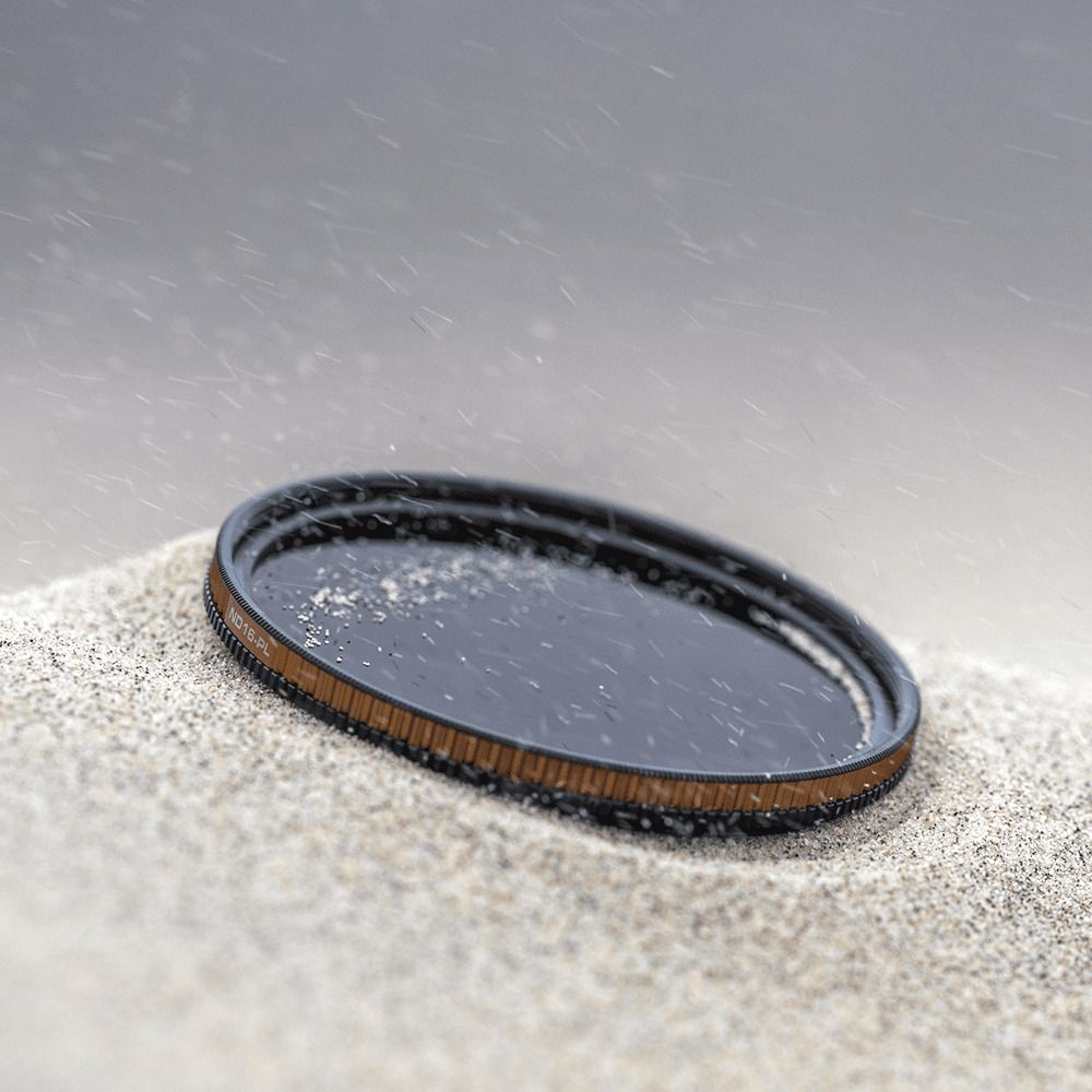 Een rond ND18-PL filter rust op zand, met regendruppels op het glas.