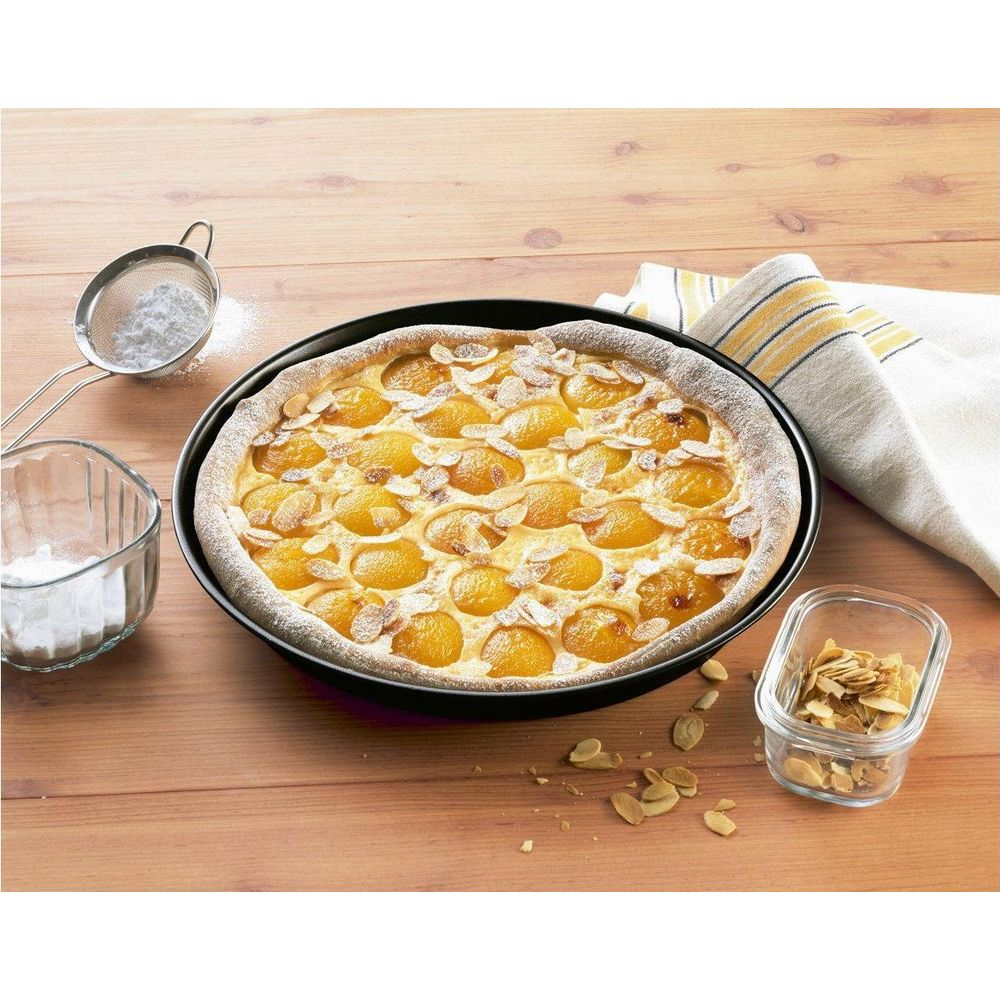 Une tarte ronde aux abricots dans un plat noir, garnie d'amandes effilées et de sucre glace.