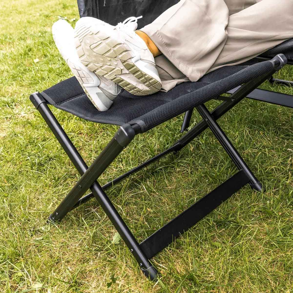 Voeten in witte sneakers op zwarte klapstoel boven groen gras.