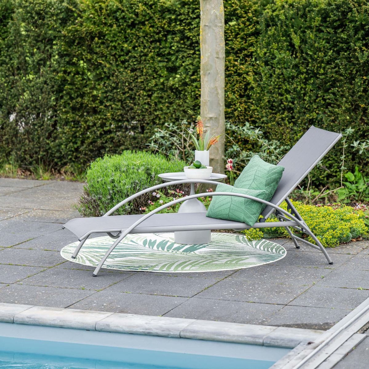 Grijze ligstoel met groene kussens op terras bij zwembad.
