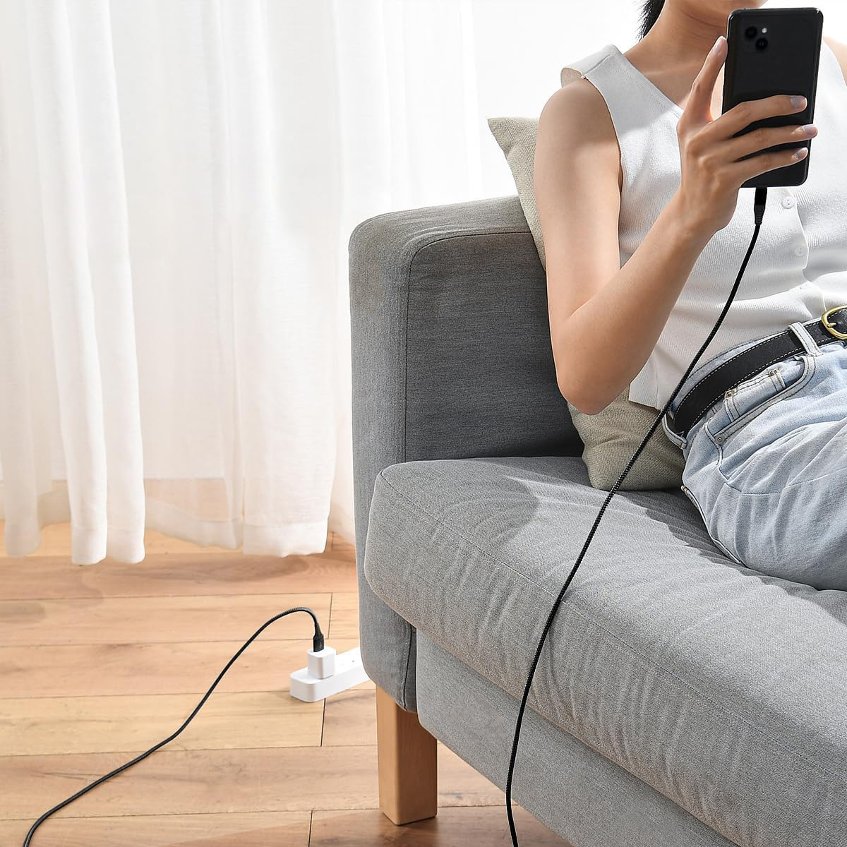 Eine Person sitzt auf einem grauen Sofa und lädt ihr Smartphone mit einem schwarzen Kabel.