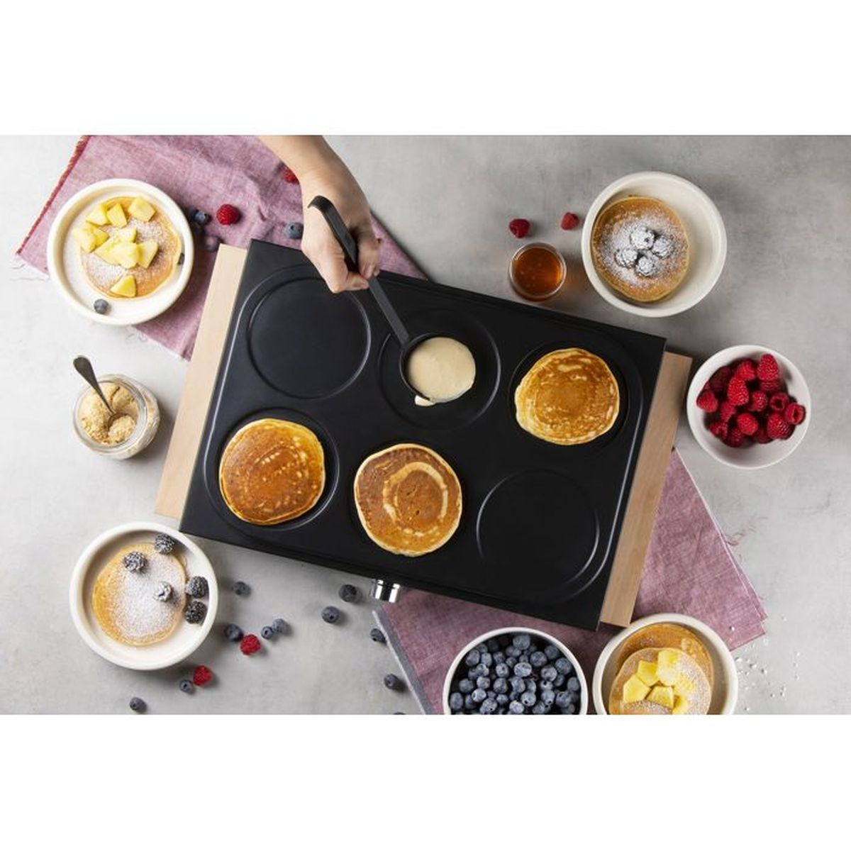 Eine Hand gießt Teig auf eine Pfannkuchenplatte mit fertigen Pfannkuchen.