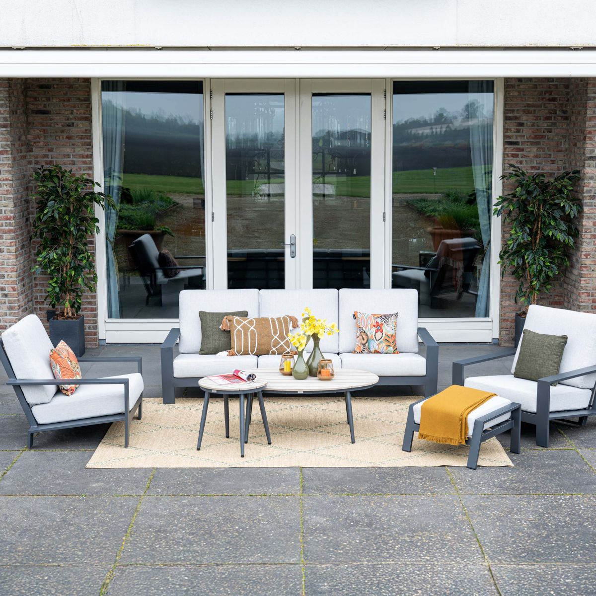 Loungeset buiten met bank, fauteuils, salontafels en vloerkleed voor glazen deuren.