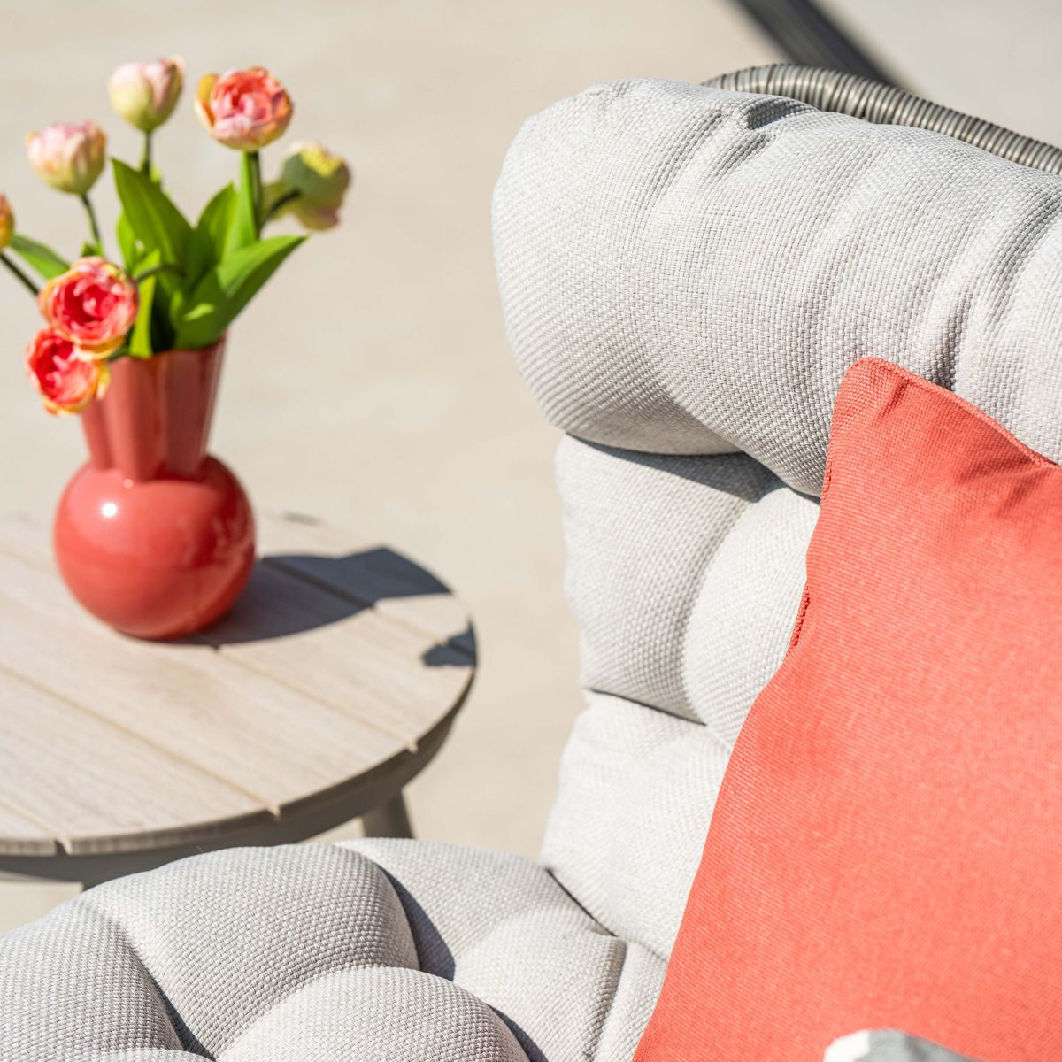 Een koraalkleurige vaas met tulpen staat op een houten tafel naast een fauteuil met rood kussen.