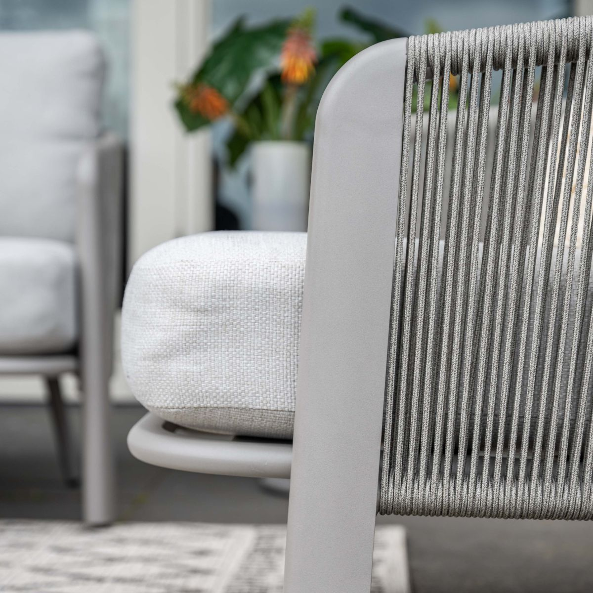 Grijze loungestoel met touwrugleuning en wit kussen, plant op achtergrond.