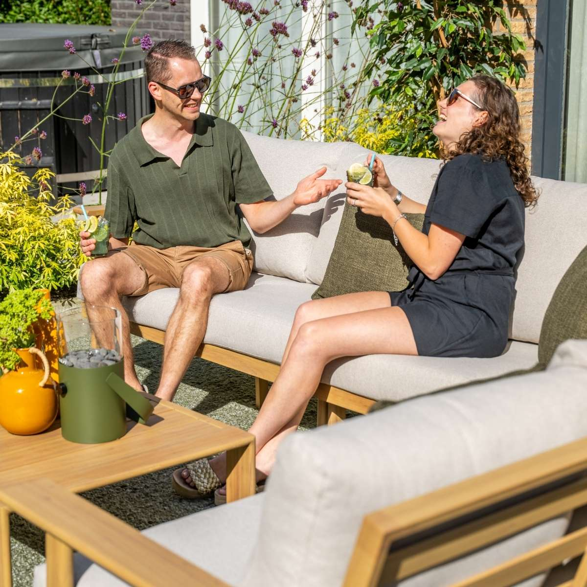 A man and woman sit on an outdoor couch, holding drinks and talking.