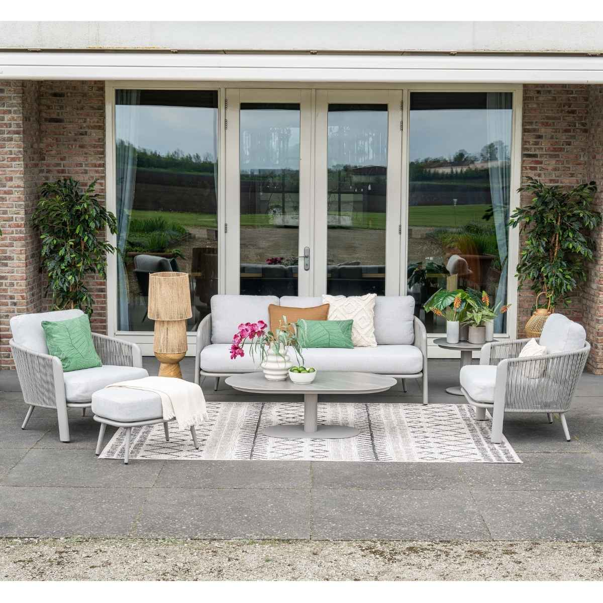 Grijze tuinmeubelen met kussens op een terras met planten en glazen deuren.