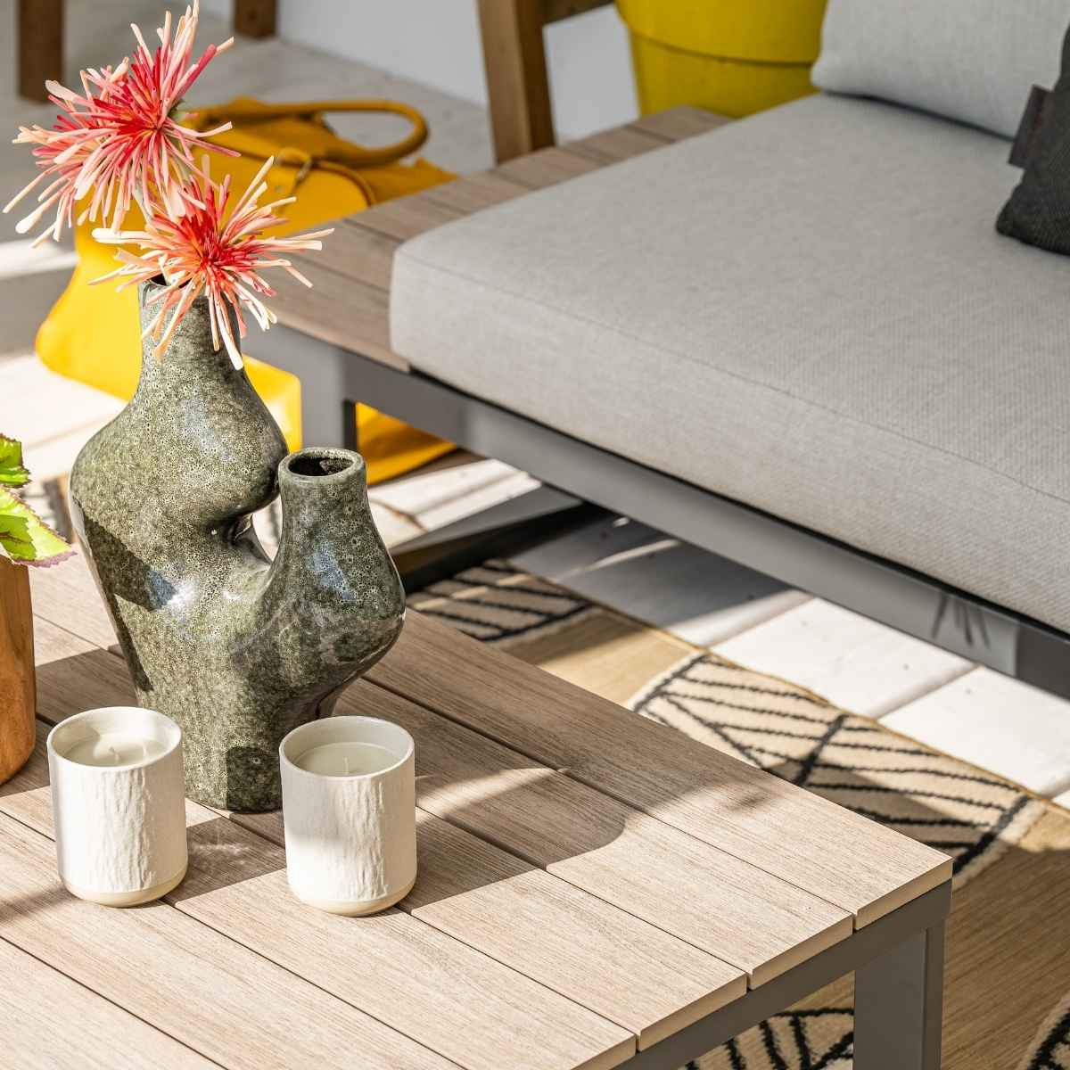 Wooden table with two candles, green vase with flowers, yellow bag, sofa, and rug.