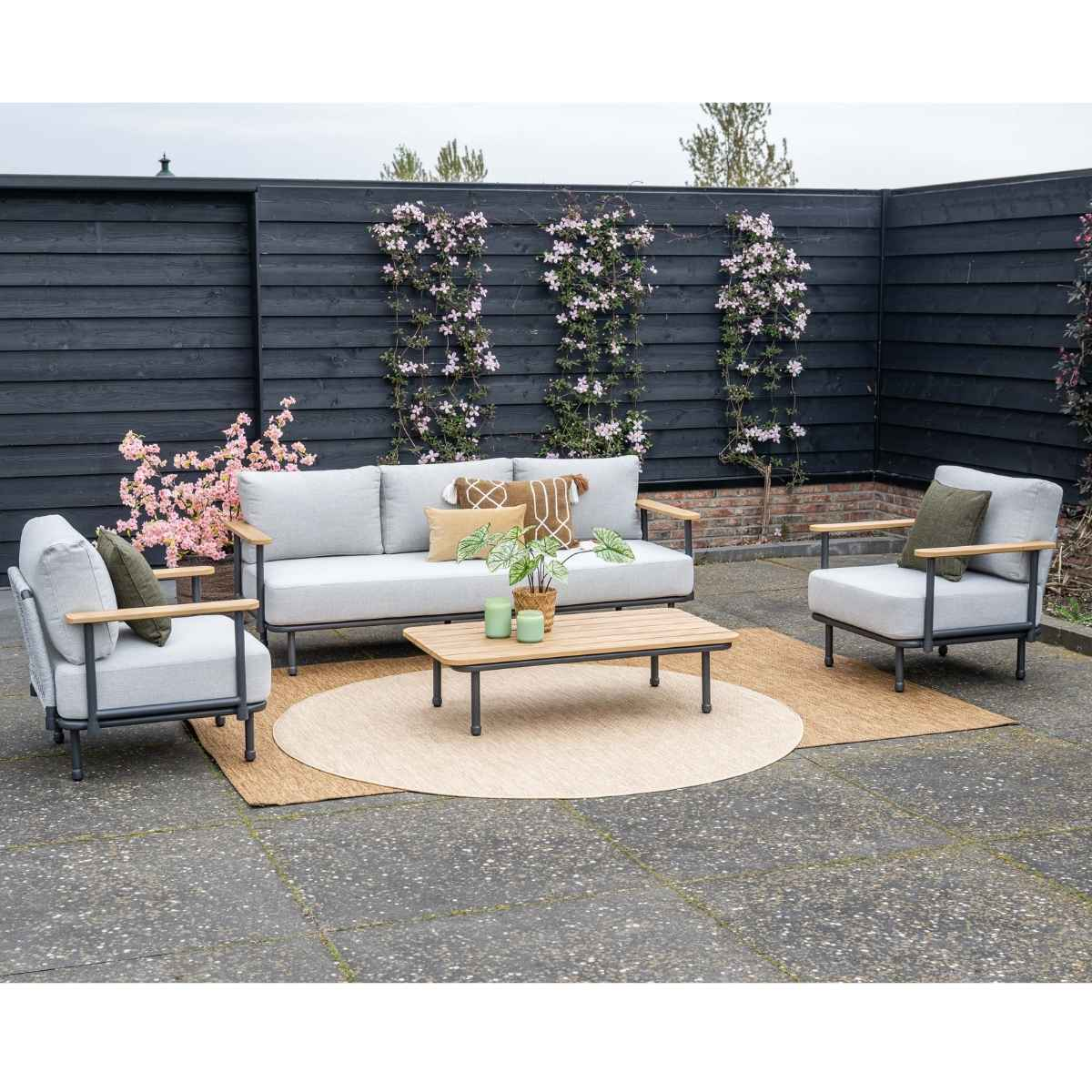 Outdoor lounge set with grey sofa, armchair, wooden table, and plants before black fence.