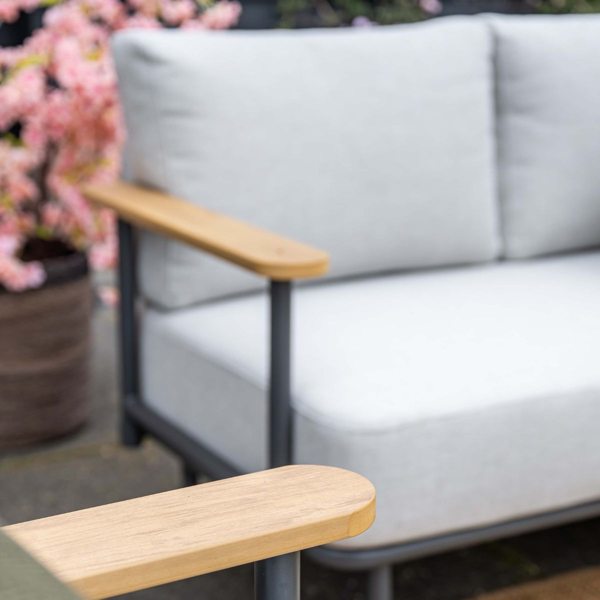A grey sofa with wooden armrests sits next to a pot of pink blossoms.