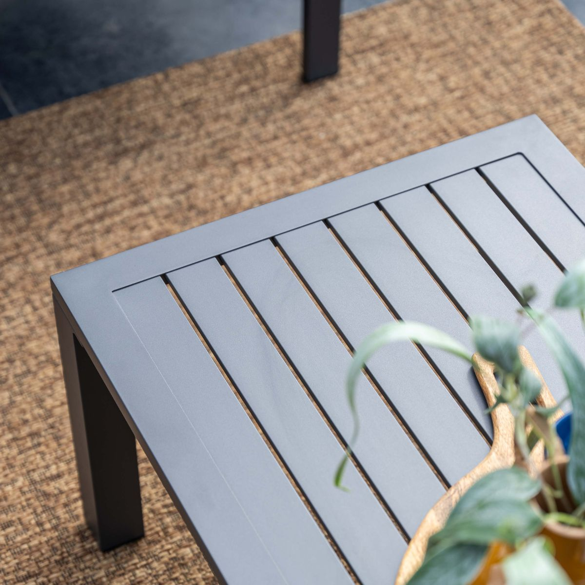 Gray slatted table on a brown rug with a potted plant in the foreground.