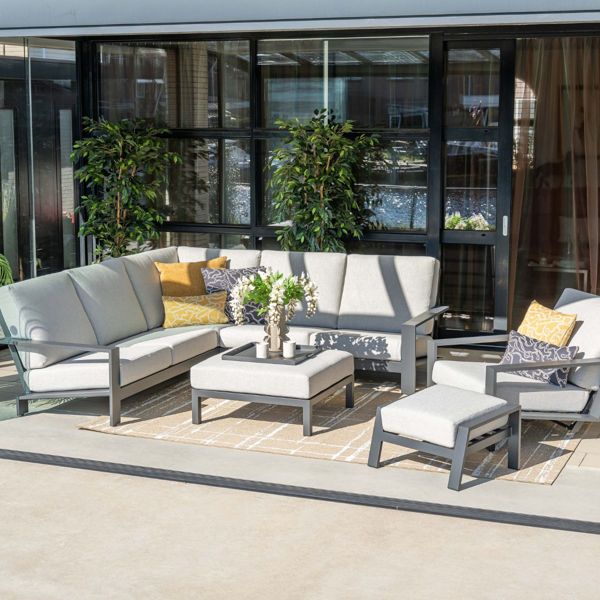 Gray sectional sofa with cushions and ottoman on rug in front of glass windows.
