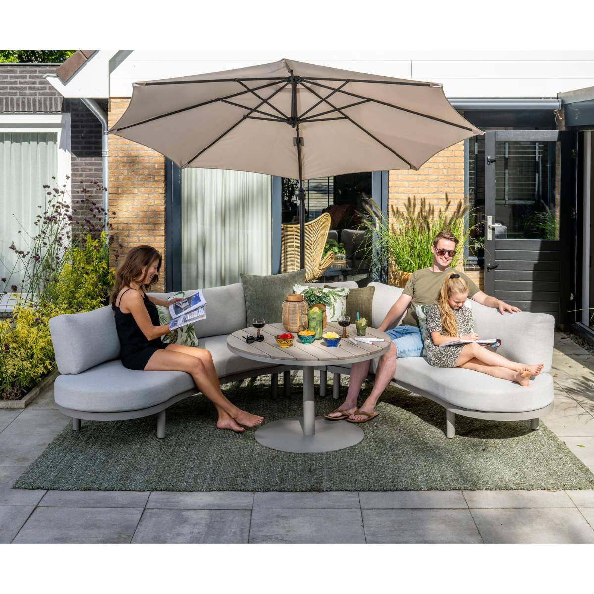 Family relaxing on outdoor sofa under umbrella with table. Woman reads, man and child draw.
