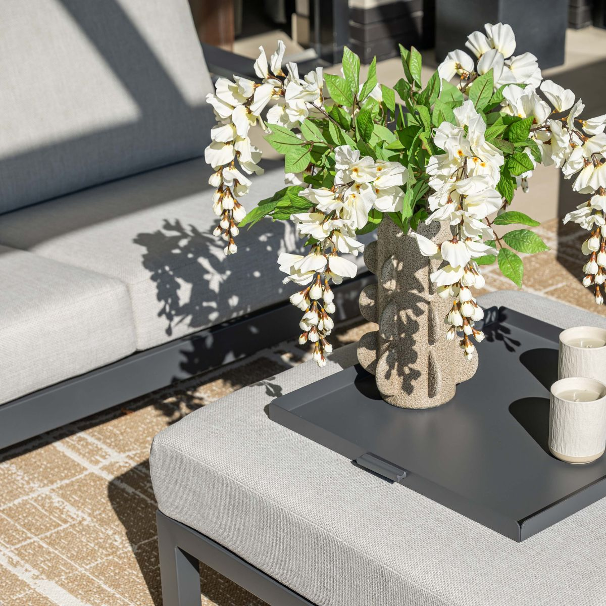 A bouquet of white flowers in a vase sits on a tray next to two candles on an ottoman.