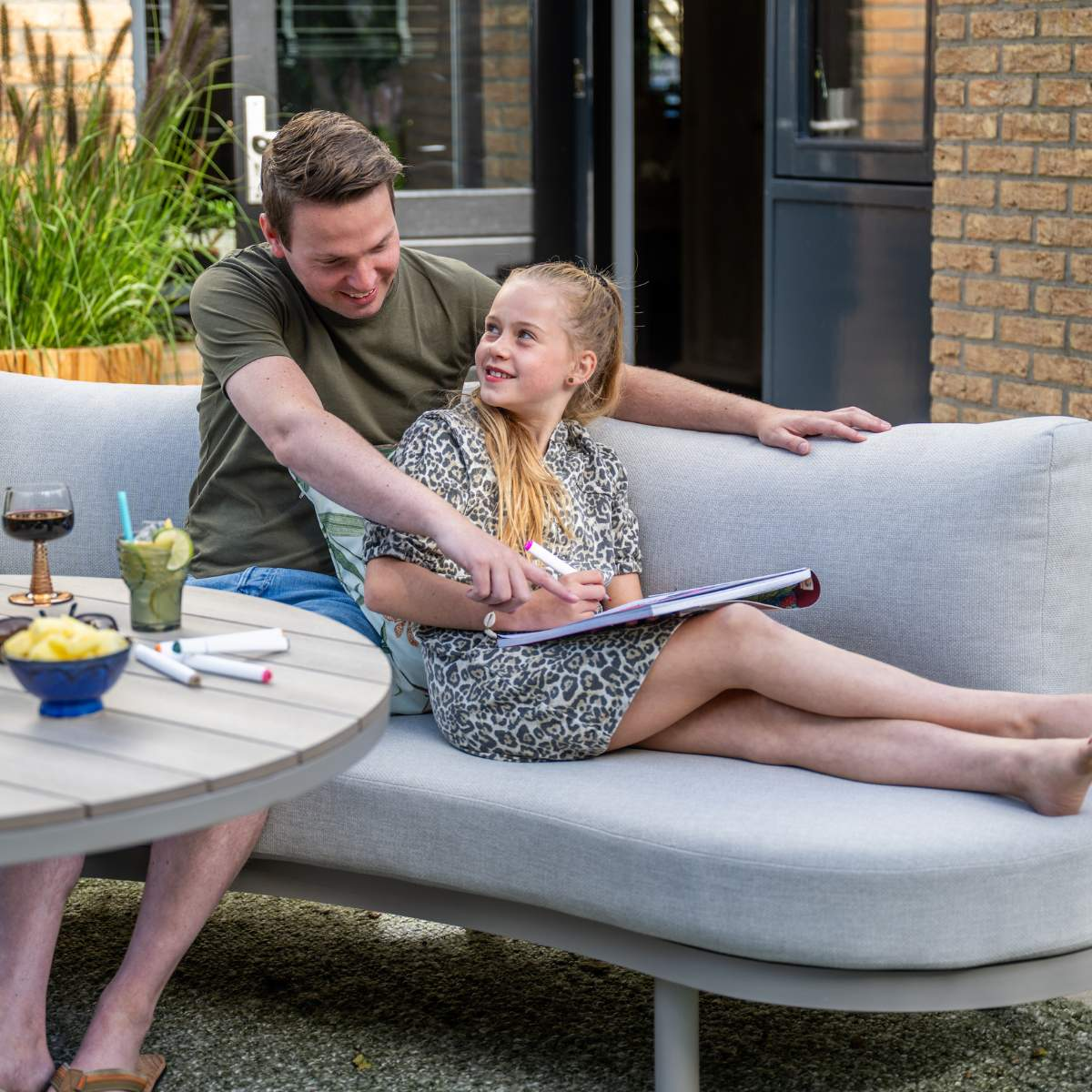 Man and girl sit on sofa, drawing outside. Drinks and snacks on table.