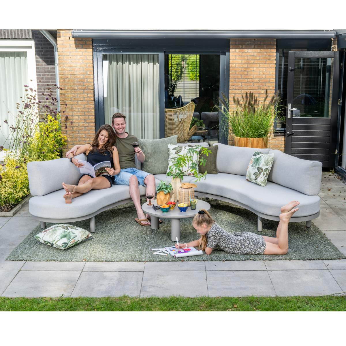 A couple relaxes on a sectional sofa while a girl draws on a rug in the garden.