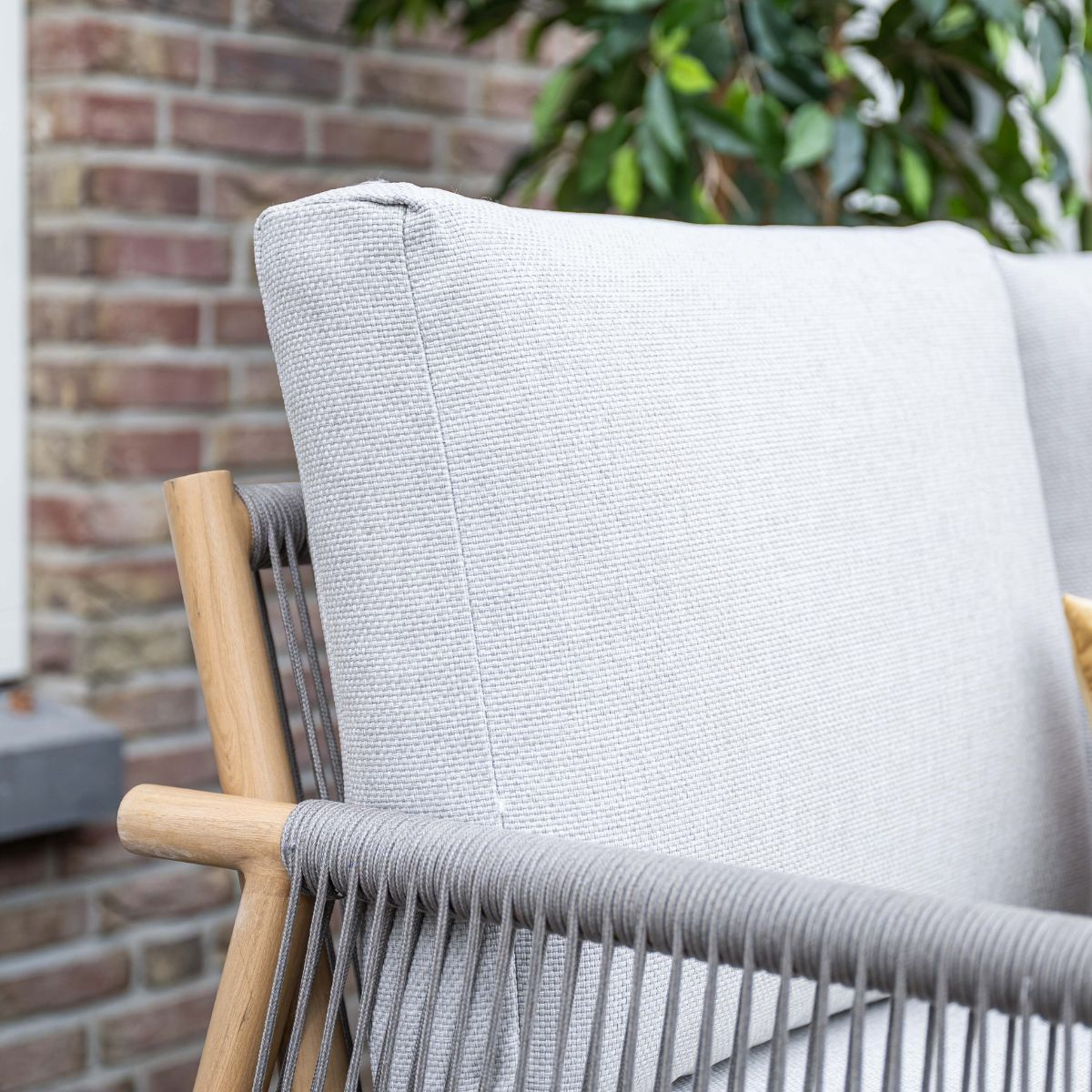 Grijze fauteuil met houten accenten en touwdetails, bakstenen muur en groene plant.
