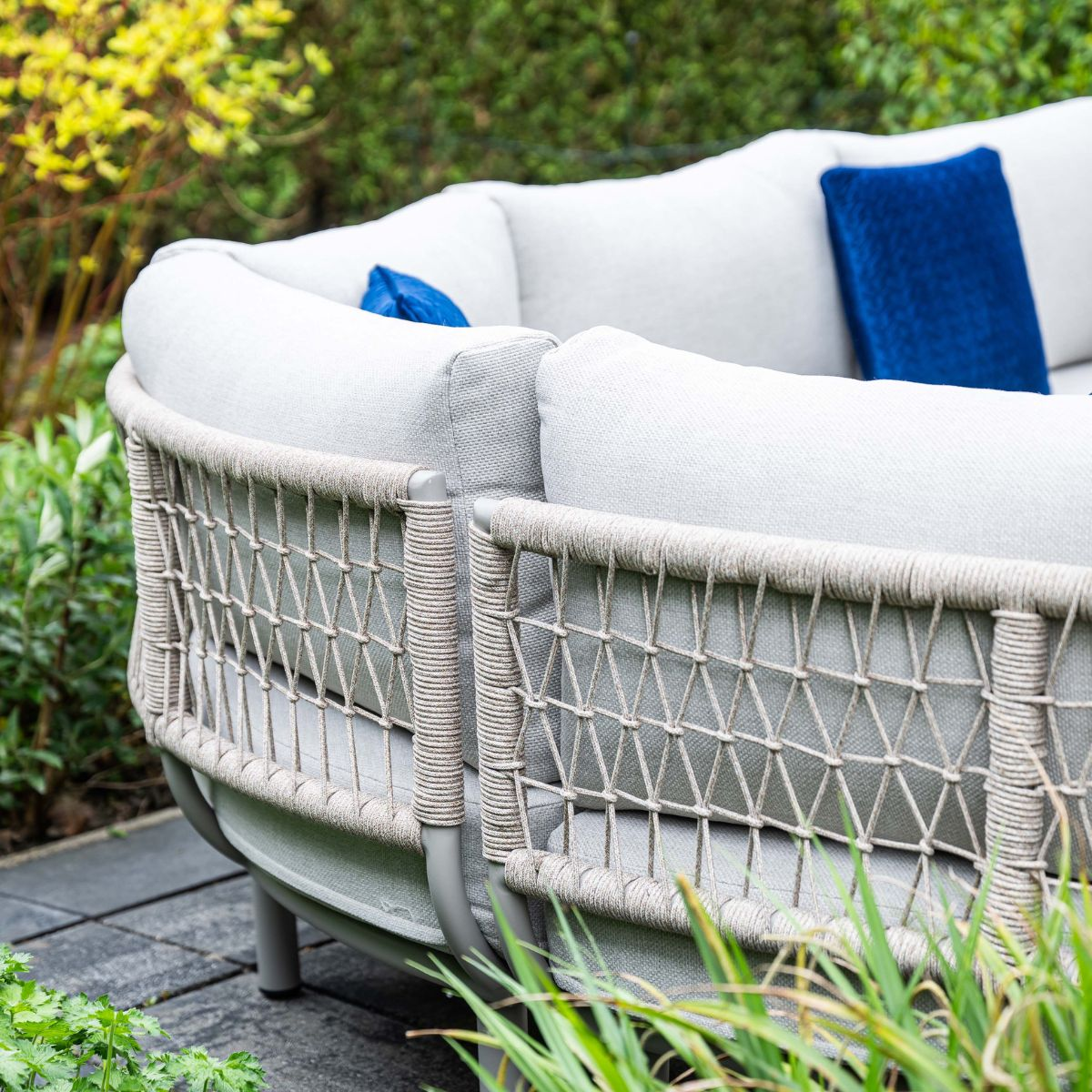 Grijze buitenbank met witte kussens en blauwe accentkussens, in tuin.