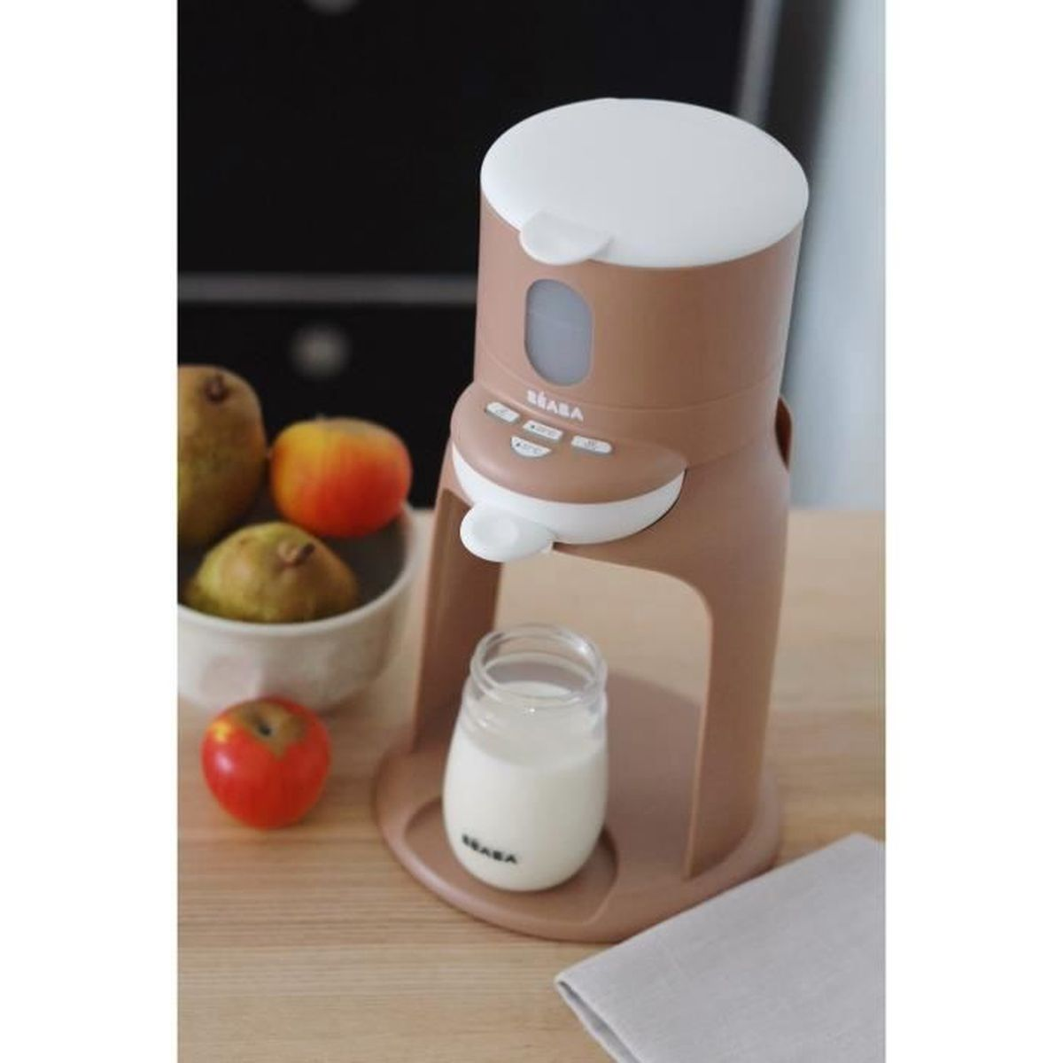Ein brauner Babykostbereiter mit weißem Deckel steht auf einem Holztisch. Daneben sind Früchte und ein Glas Milch.