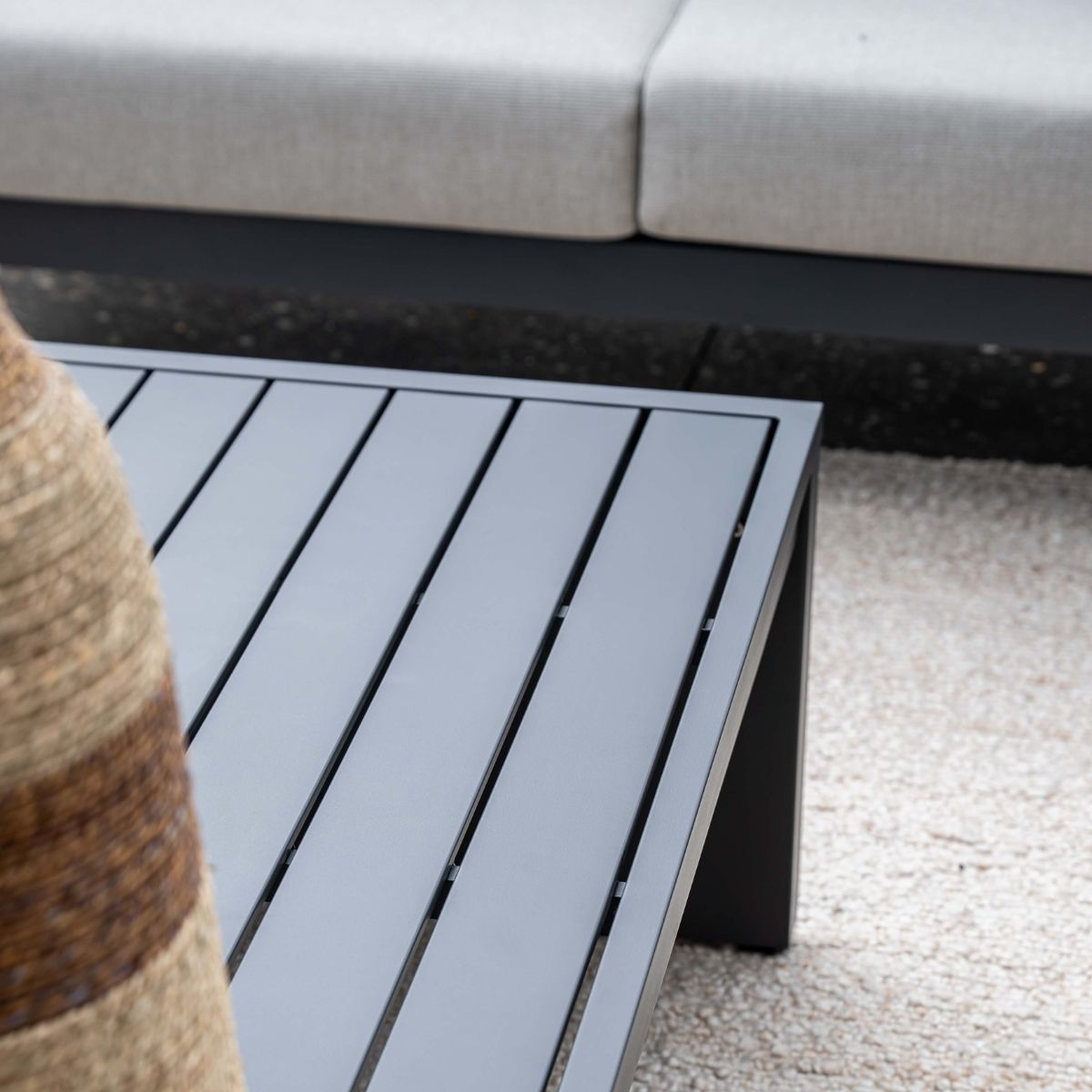 Gray slatted coffee table on light gravel with gray sofa in background.