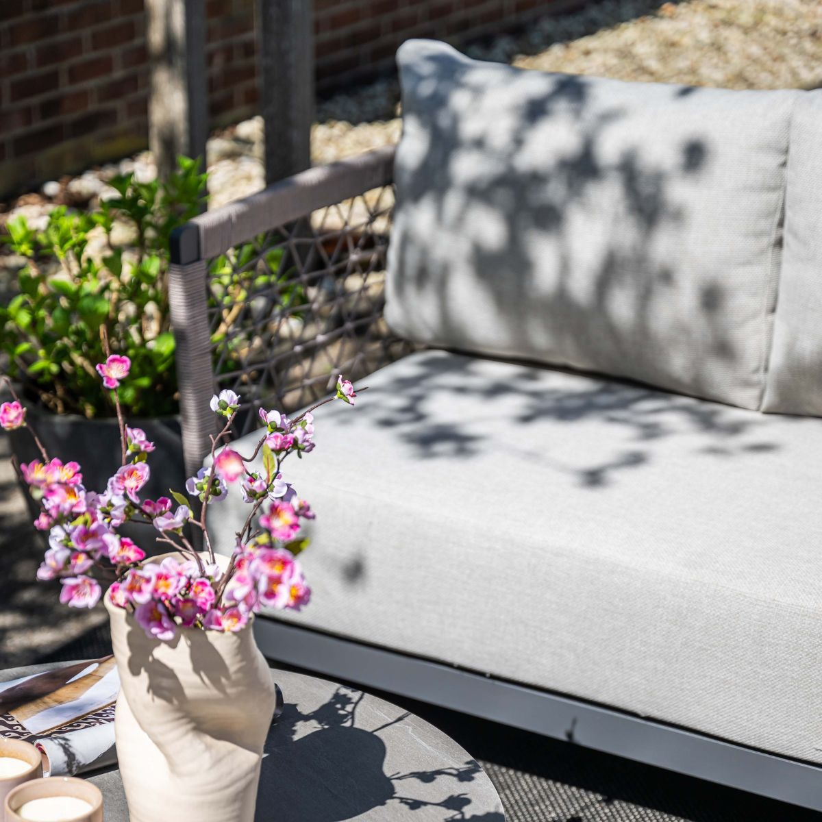A vase with pink blossoms on a table next to a light grey outdoor sofa.