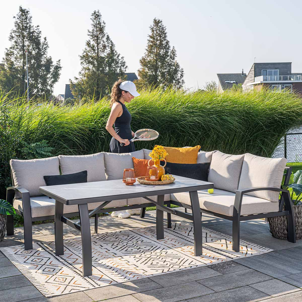 Vrouw met racket naast tuinmeubilair met tafel en kussens.