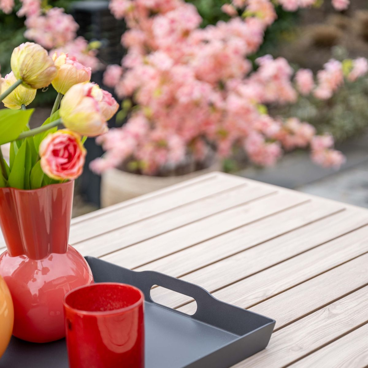 Een rode vaas met tulpen op een houten tafel, ernaast een rood glas en een dienblad.