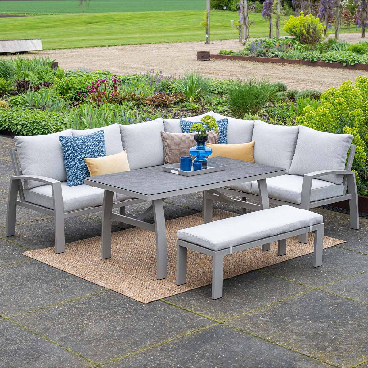 Grijze lounge set met tafel en bank op een terras met bloembedden.
