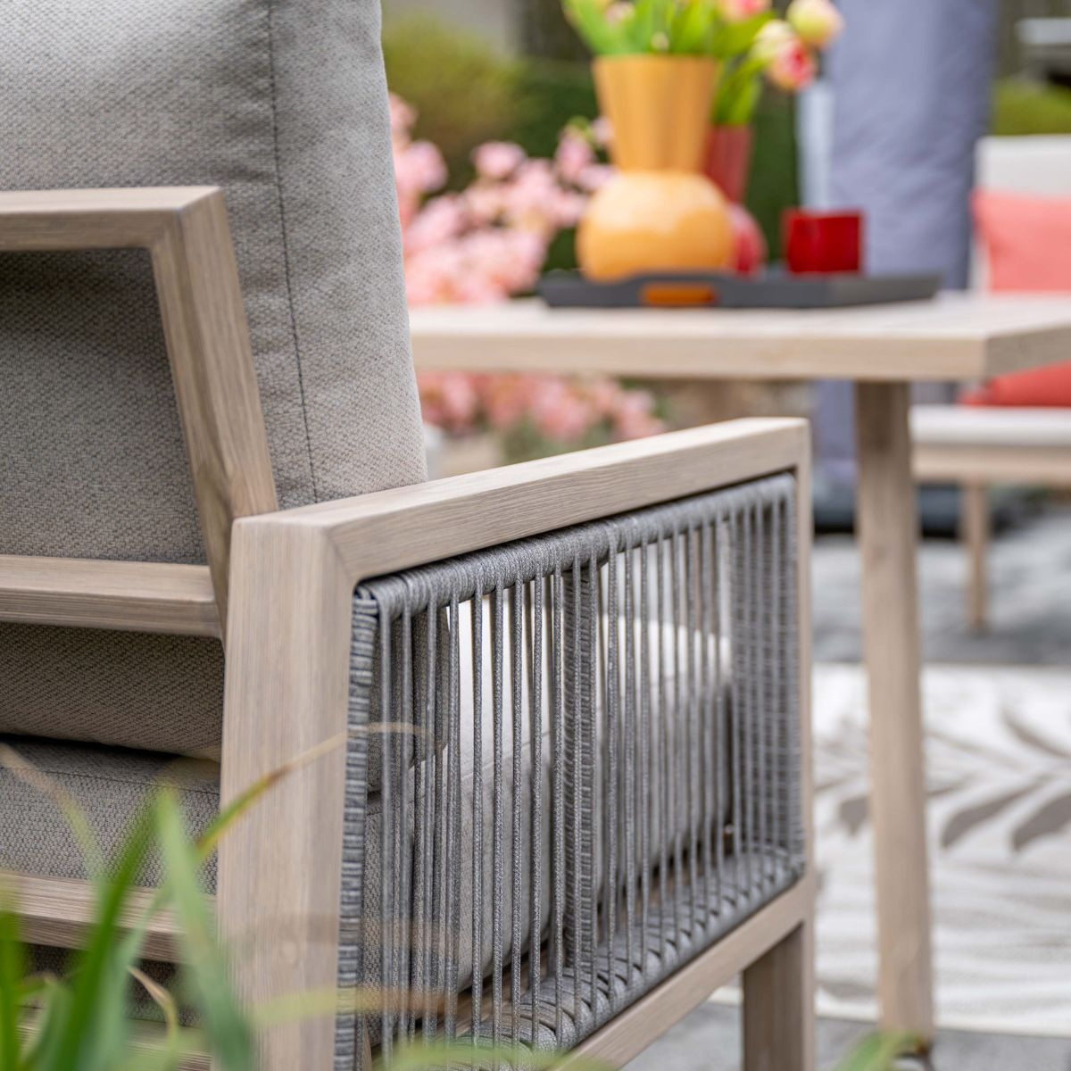 Moderne houten tuinstoel met grijze touwvlecht, kussens en tafel op achtergrond.