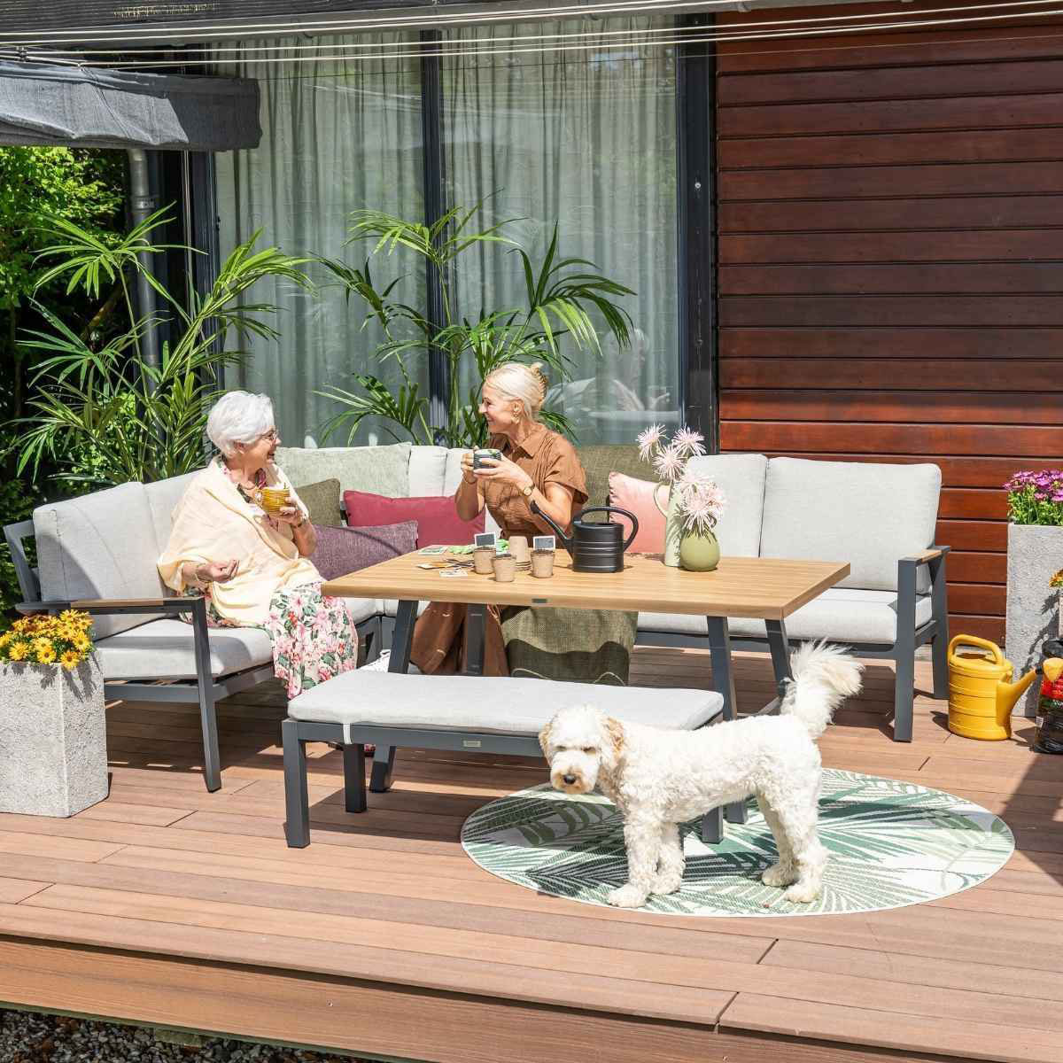 Twee vrouwen en een hond op een terras met tuinmeubelen en planten.