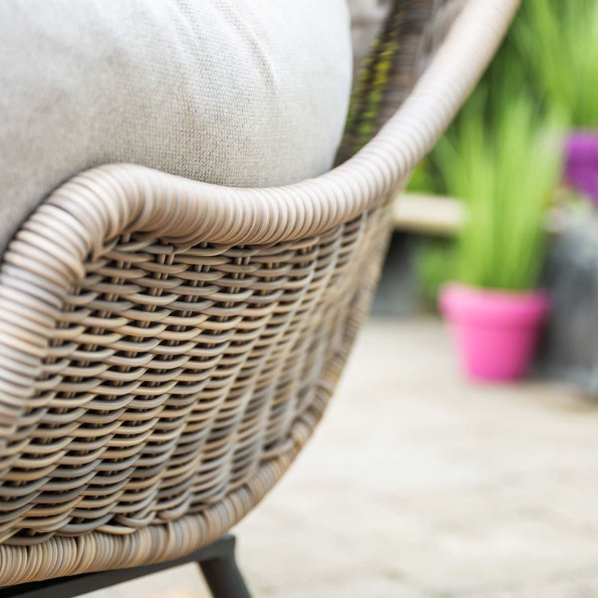 Een rotan stoel met grijs kussen buiten, met planten op de achtergrond.
