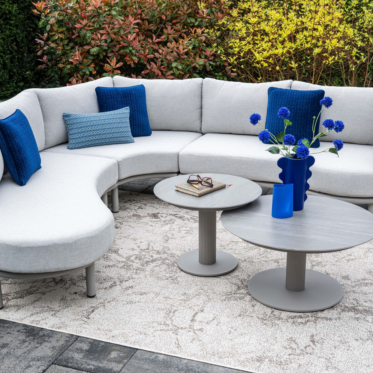 A grey outdoor sectional sofa with blue cushions, two coffee tables, and flowers.