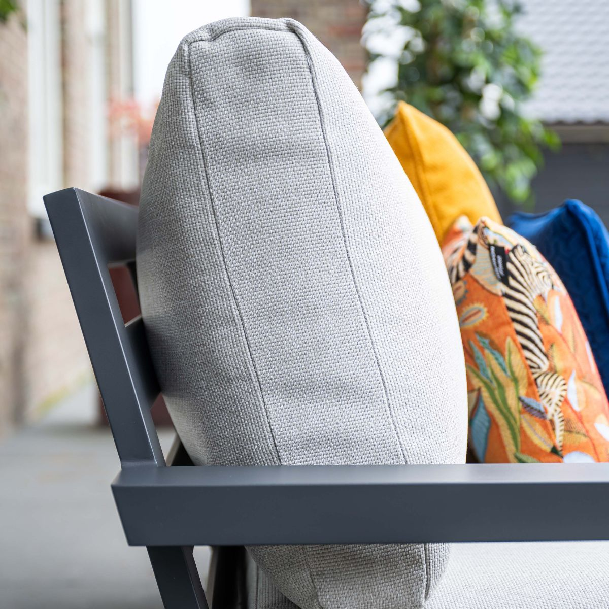 Een grijs kussen op een donker metalen frame, met gele, oranje en blauwe kussens erachter.