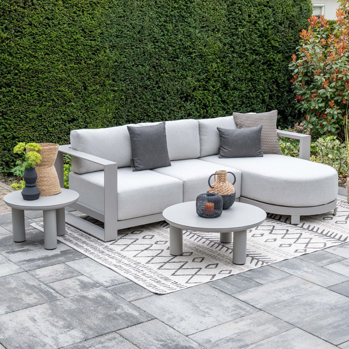 A grey sectional sofa with cushions and side tables on a paved patio.