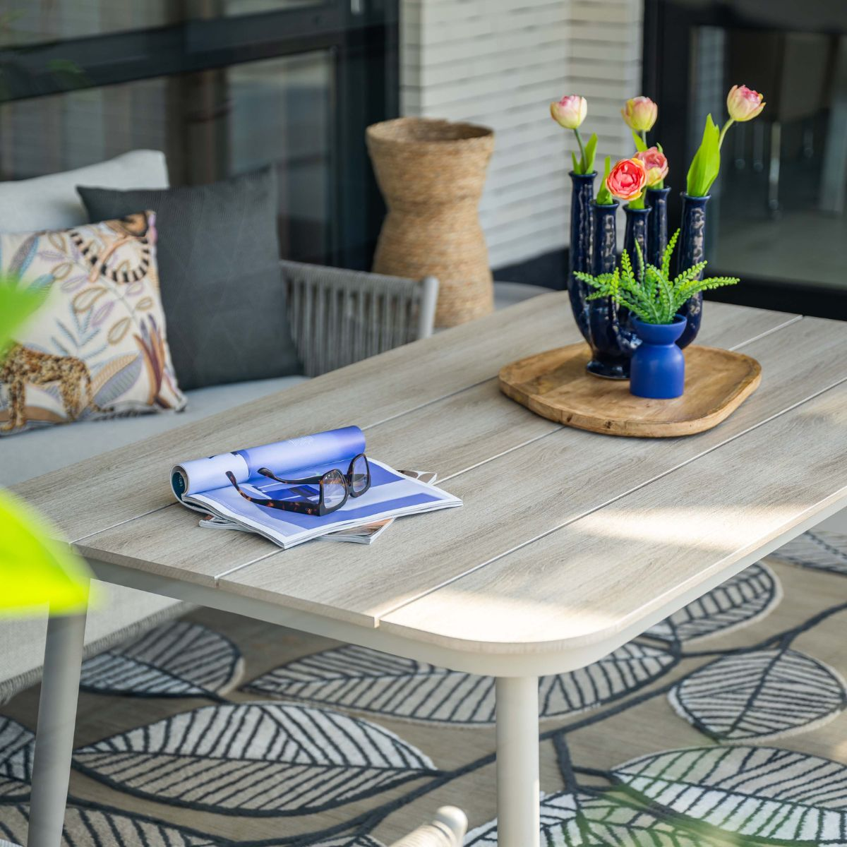 Houten tafel met tijdschriften, bril, blauwe vazen met bloemen en groene plant.