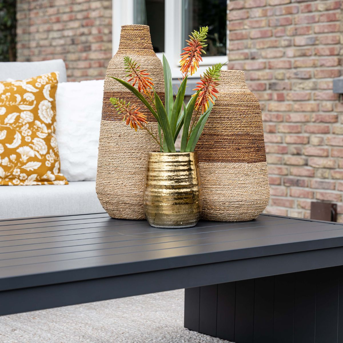 Two woven vases with flowers in a gold vase sit on a table.