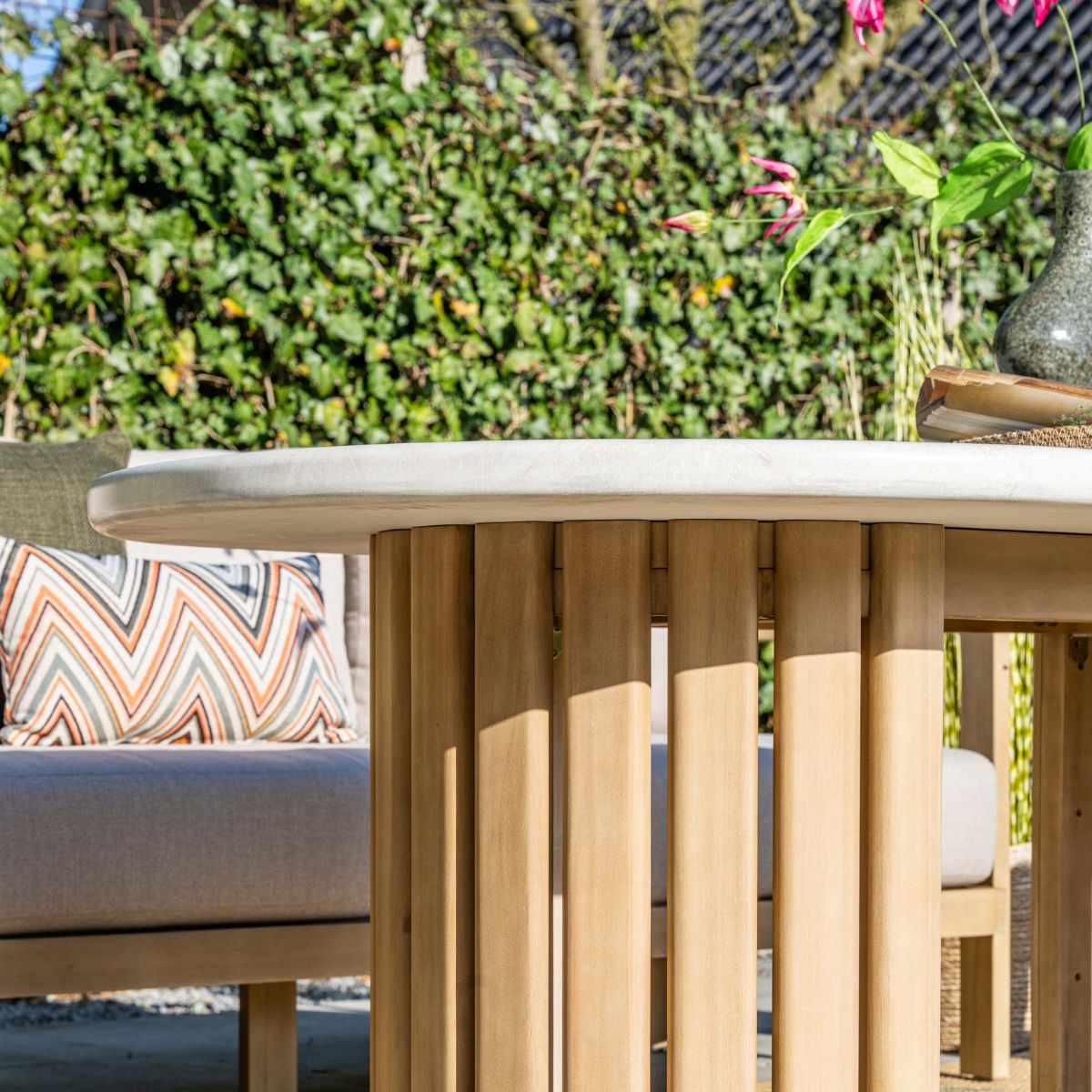 Een ronde betonnen en houten tafel in een tuin met kussens op een bank.