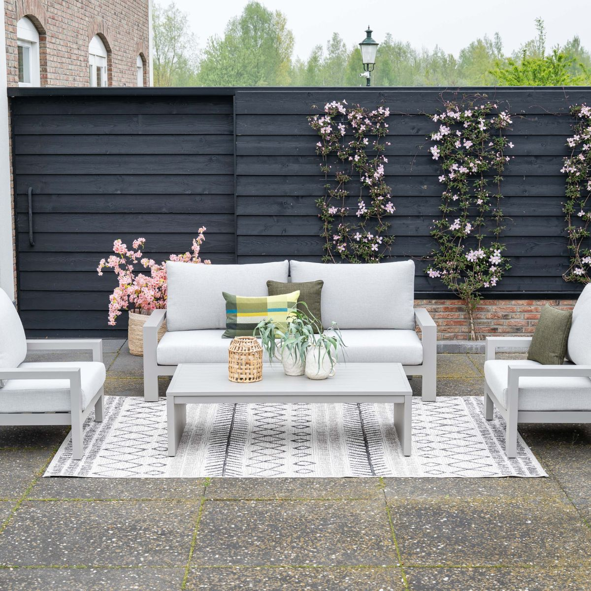 A grey outdoor sofa and chairs with cushions on a patio with a rug and plants.