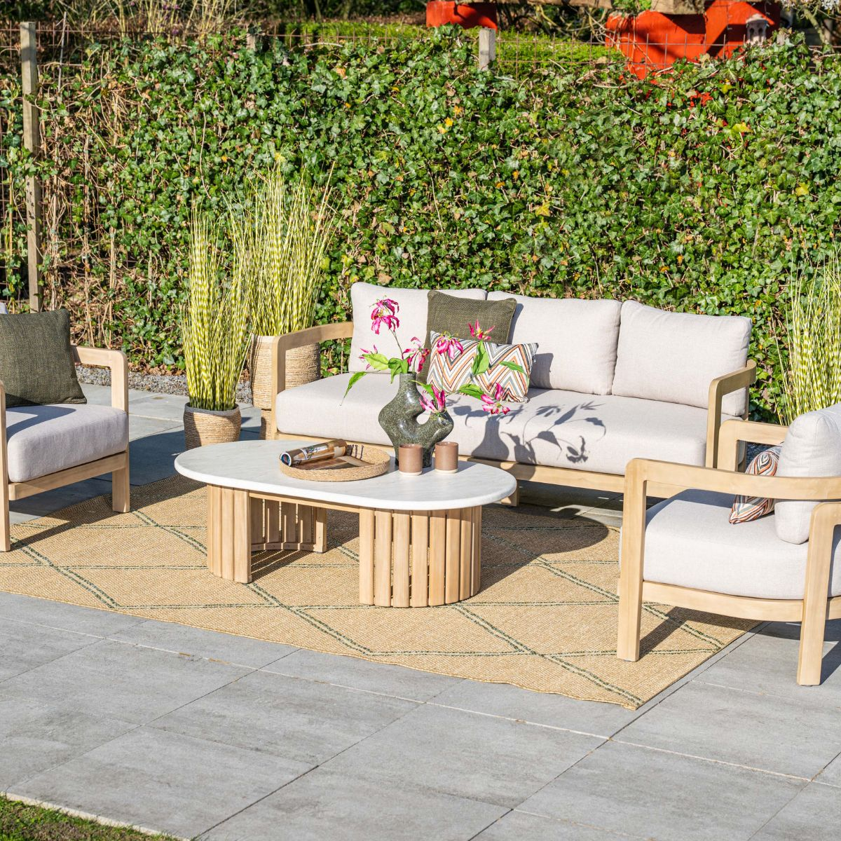 Outdoor lounge set with sofa, armchairs, coffee table, and rug in front of hedge.