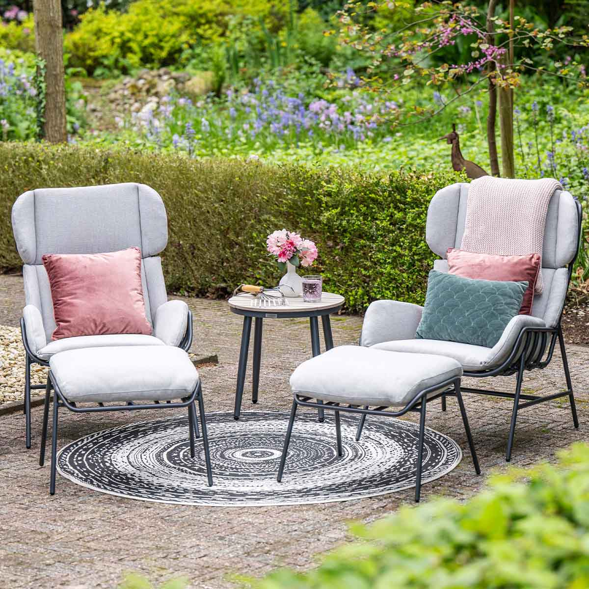 Twee grijze fauteuils met poefs en kussens op een terras met bloemen.